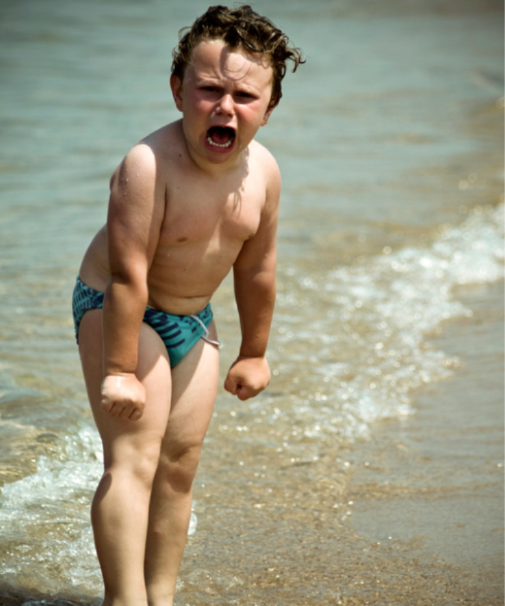 Upset toddler boy in swimsuit throwing a tantrum at the beach, fists clenched and face angry