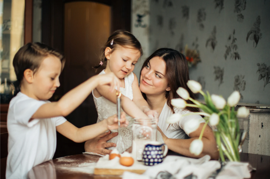 Candid family moment with mom and kids baking together in the kitchen. Flour is on the table and their faces, with the kids having fun playing with kitchen tools