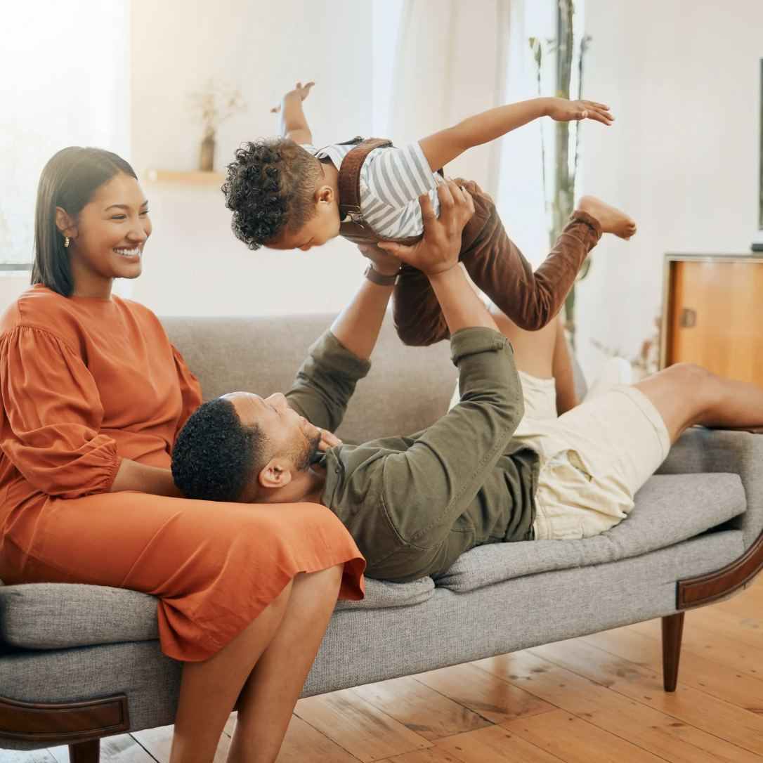 Happy mother sits on a couch watching her partner lift their toddler, creating a moment of joy in a calm home environment.
