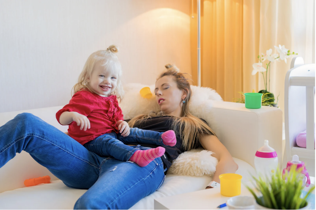 Exhausted mother asleep on the couch with toys around her and a happy toddler sitting on top of her chest, symbolizing sleep deprivation in parenting.