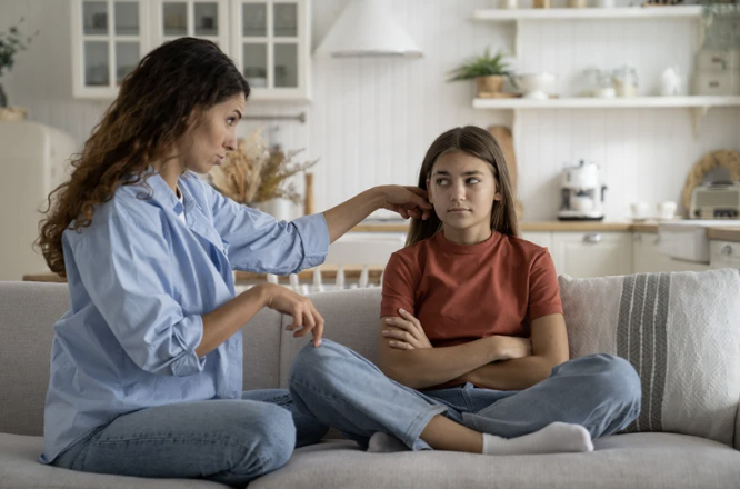 Mother setting firm but compassionate parenting boundaries with skeptical preteen daughter on couch.