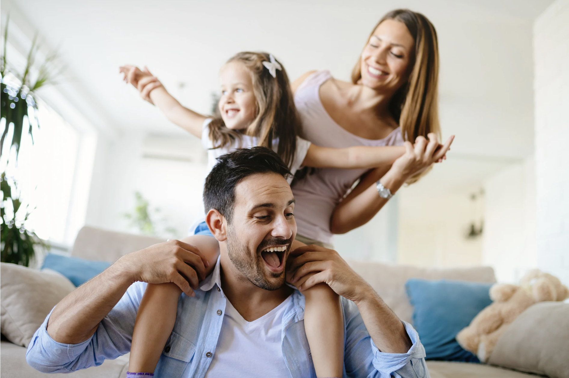 Happy family moment in a bright living room — dad playing with his daughter on his shoulders while mom joins in, all smiling and enjoying quality time together.