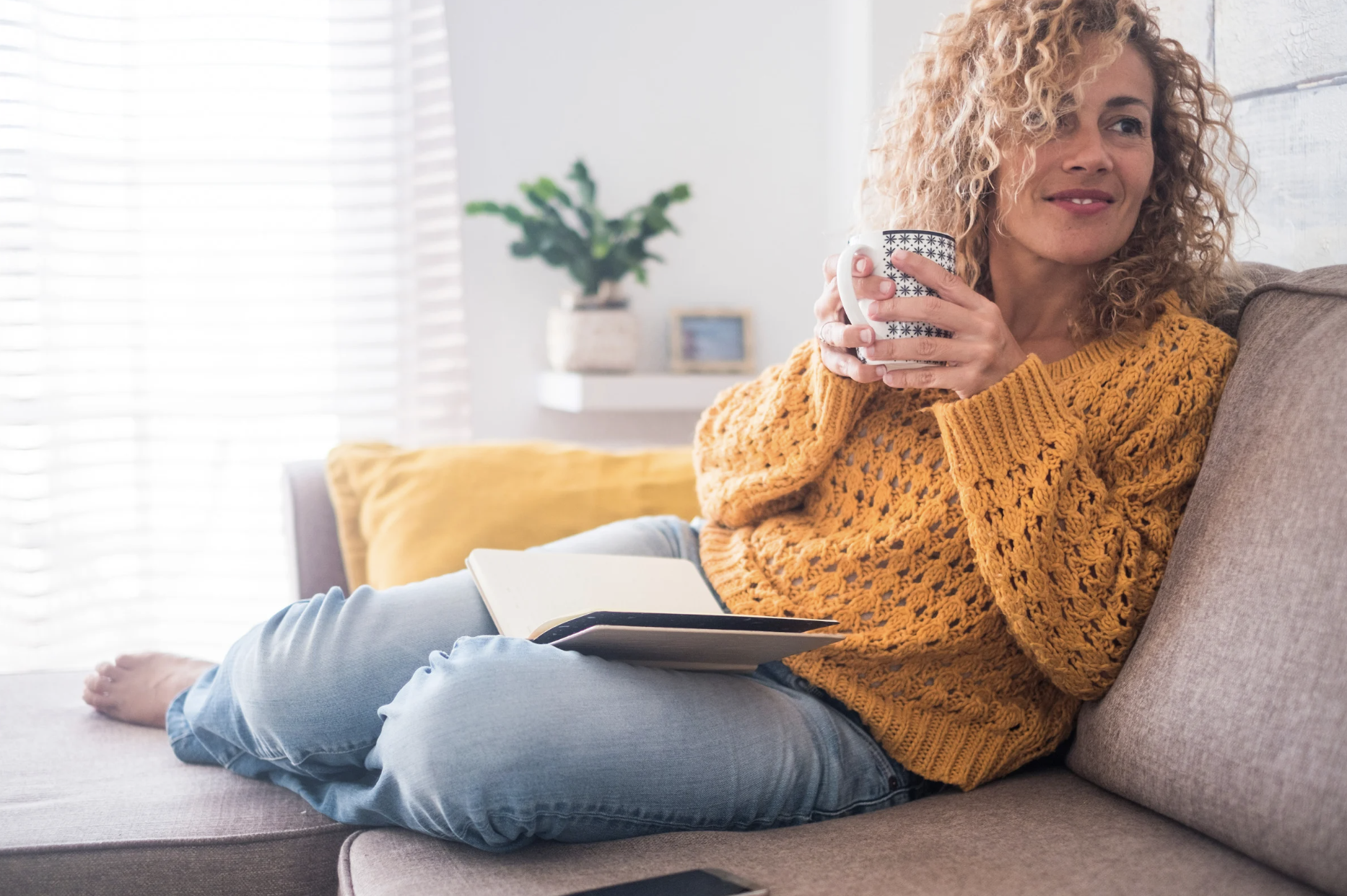 Woman relaxing on a couch with a mug and open journal, embracing a moment of self-care in a calm, minimalist setting.