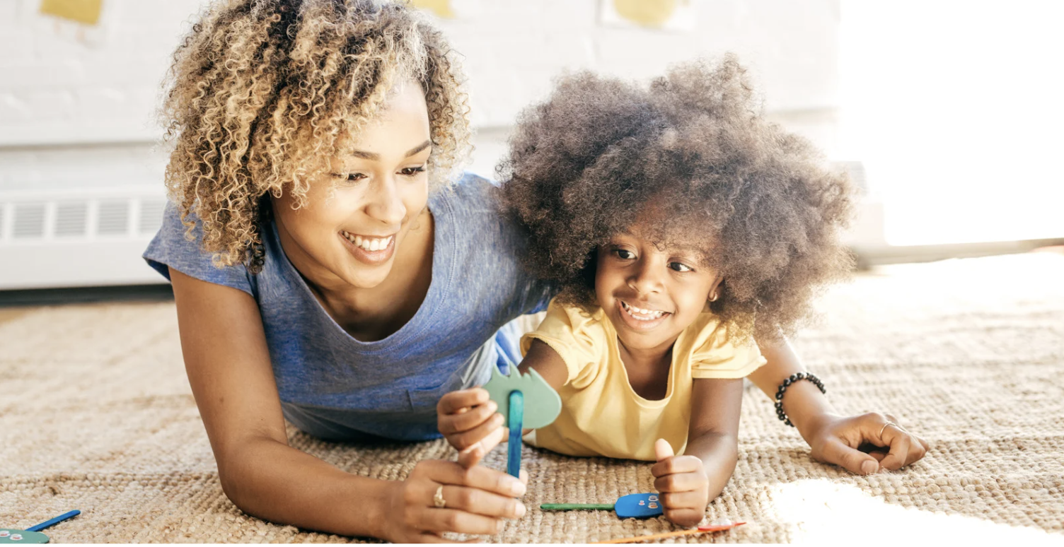 Mom and toddler lying on the floor playing with popsicle stick puppets, sharing a joyful, carefree moment together