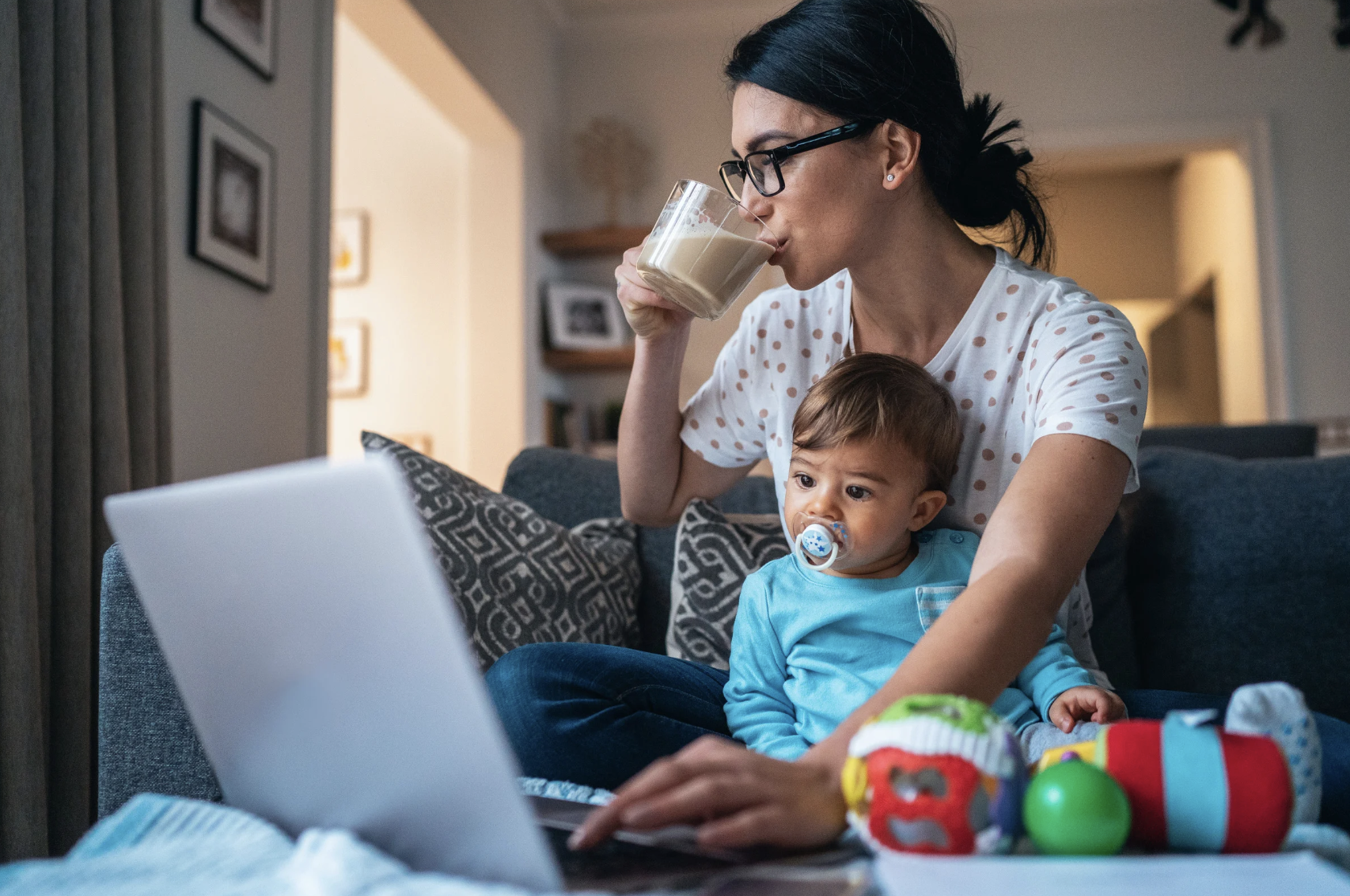 Young mom with her hair pulled back, holding a coffee mug and typing on a laptop with her baby on her lap and toys scattered in the foreground — a relatable snapshot of a multitasking mom in need of stress management.
