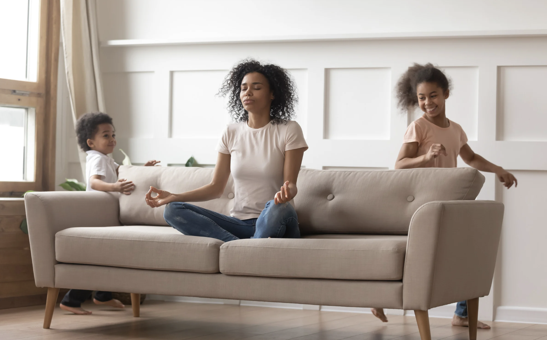 Mom sitting in a lotus position trying to meditate on the couch while two young kids run wild around her, capturing the chaos and stress of motherhood.