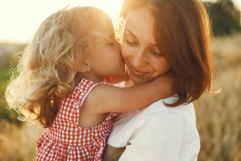 Mother holding toddler daughter outdoors in a peaceful embrace as sunlight shines between their faces — a quiet moment of connection that shows parenting is hard, but full of love.
