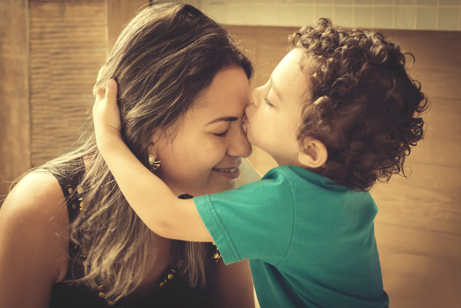 A tender moment between a tired mom and her curly-haired toddler as he kisses her forehead, capturing the beauty and exhaustion of parenting when it feels impossible.