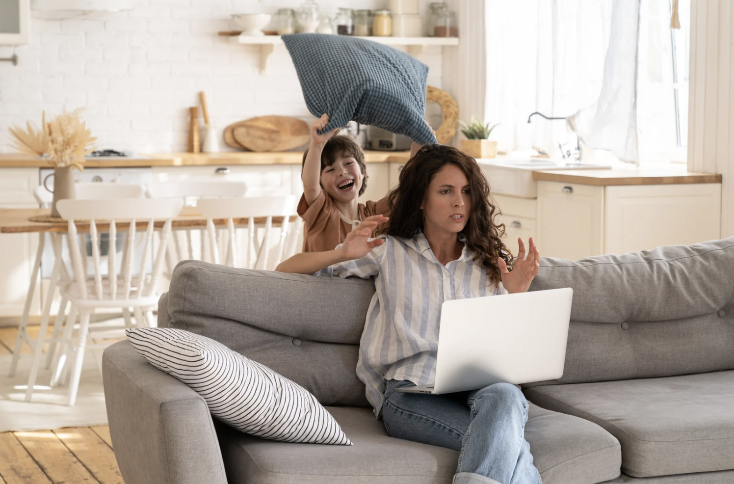 Stressed mom tries to work on laptop while toddler raises a pillow to hit her, illustrating real-life mom burnout symptoms.